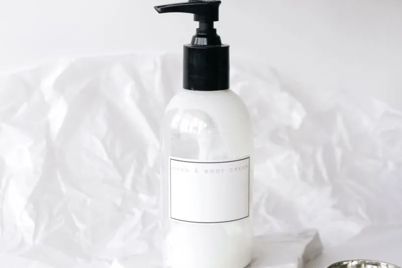 Flacon-pompe blanc sans marque, étiqueté « Hand & Body Cream », devant un papier de soie froissé