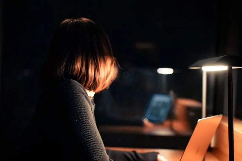 Femme vue de dos, seule devant un ordinateur portable dans un bureau plongé dans le noir, éclairée par une lampe