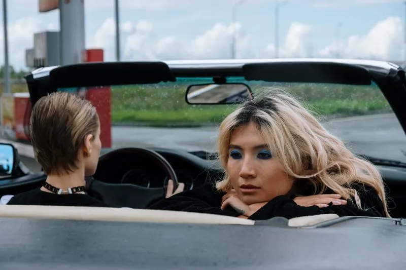 Deux femmes dans un cabriolet décapoté, la conductrice vue de dos, la passagère penchée sur le dossier, le menton posé sur ses bras