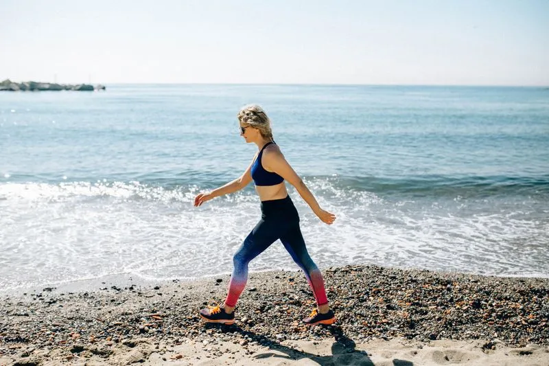 Femme en tenue de sport qui marche d'un bon pas sur une plage de galets, au bord de l'eau