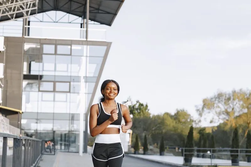Jeune femme souriante en brassière de sport et short noirs qui court sur une promenade, devant un bâtiment vitré, dans une lumière douce