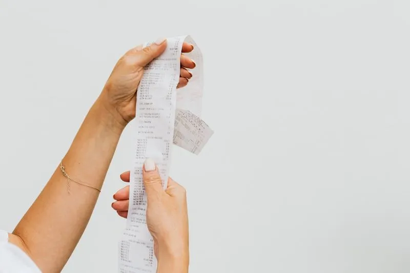 Mains tenant un long ticket de caisse déroulé devant un mur blanc