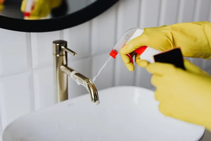 Mains gantées de caoutchouc jaune vaporisant un produit sur un robinet doré au-dessus d'une vasque blanche