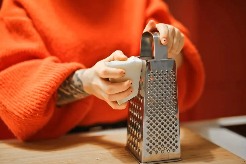 Mains râpant un pain de savon blanc sur une râpe en inox, sur un plan de travail en bois