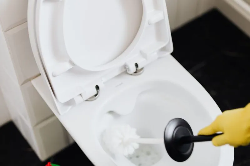 Main gantée de jaune frottant l'intérieur d'une cuvette de WC blanche avec une brosse, abattant relevé