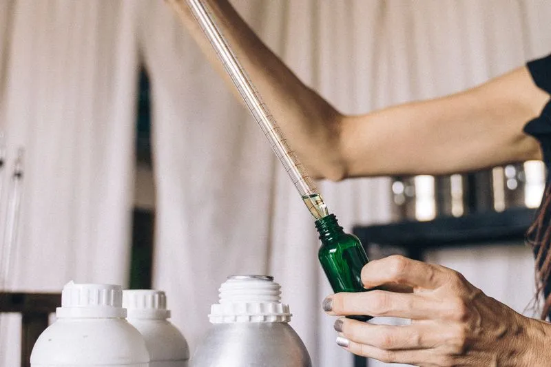 Main dosant un liquide à la pipette graduée dans un petit flacon en verre vert, devant des bidons blancs et métalliques
