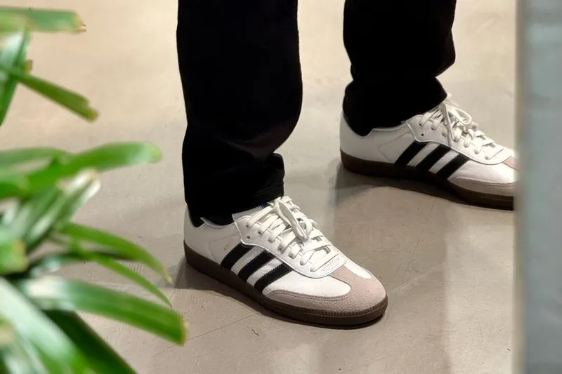 Pieds en jean noir chaussés d'Adidas Samba blanches à trois bandes noires, bout en daim beige et semelle gomme