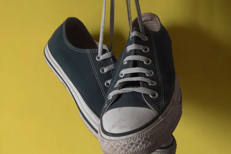 Paire de Converse basses en toile bleu nuit suspendues par leurs lacets devant un mur jaune