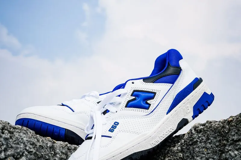 Paire de New Balance 550 en cuir blanc et bleu roi posée sur un rocher, sous un ciel nuageux