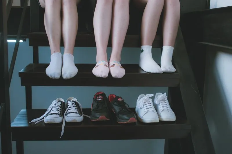 Trois paires de sneakers alignées sur une marche d'escalier en bois sombre, sous les pieds en chaussettes de trois personnes assises