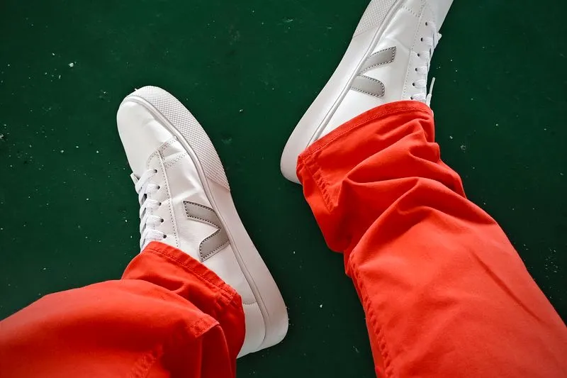 Pieds chaussés de Veja blanches en cuir au V gris, avec un pantalon rouge, sur un sol vert