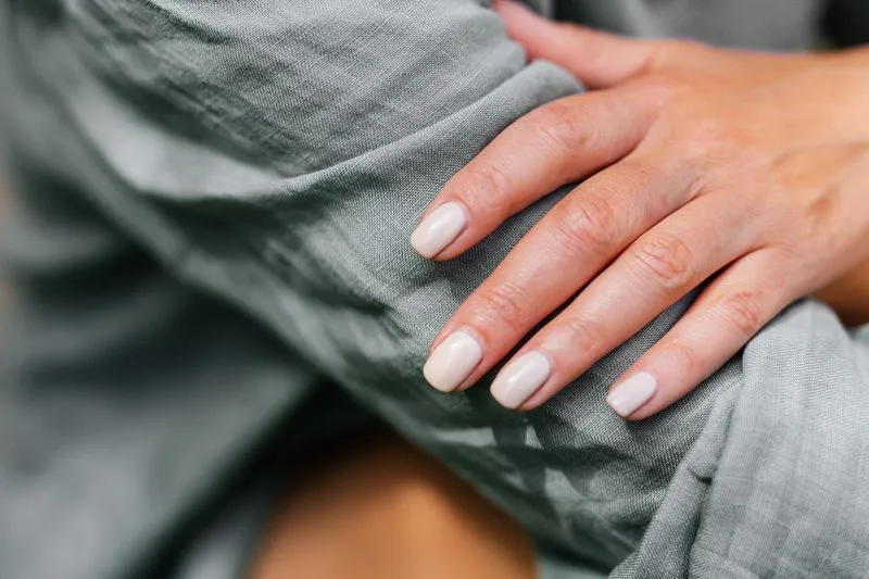 Main aux ongles courts et arrondis, vernis nude laiteux, posée sur une étoffe en lin vert-de-gris