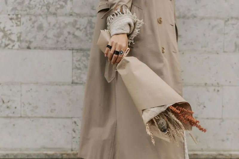 Trench beige porté sur un pull en maille crème, main baguée tenant un bouquet de fleurs séchées dans du papier kraft