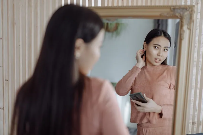 Jeune femme ajustant sa boucle d'oreille face à un miroir, téléphone à la main