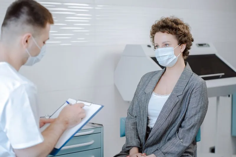 Patiente assise face à un médecin qui prend des notes sur un porte-bloc, tous deux masqués