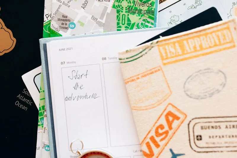 Agenda ouvert sur les mots « Start the adventure », posé sur un plan de ville, à côté d'un carnet couvert de tampons de visa et d'une boussole