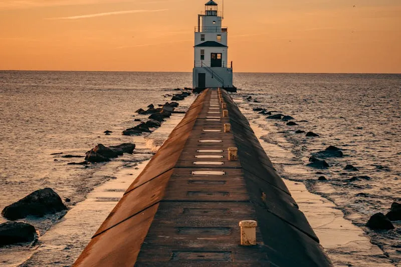 Phare blanc au bout d'une longue jetée de béton, au coucher du soleil sur une eau calme