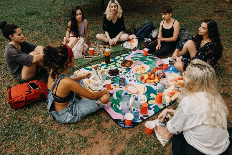 Groupe de copines assises autour d'un pique-nique partagé sur l'herbe, sacs à dos posés à côté