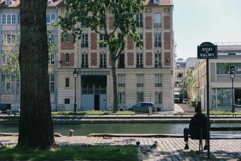Canal Saint-Martin vu depuis le quai pavé : plaque « Quai de Valmy », arbres et immeuble de brique et de pierre sur l'autre rive