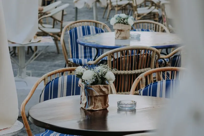Terrasse de café aux fauteuils en rotin garnis de coussins rayés bleu et blanc, petits bouquets sur les tables rondes