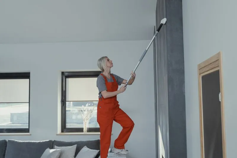 Femme en salopette montée sur un marchepied qui nettoie le haut d'un mur avec un balai à manche télescopique