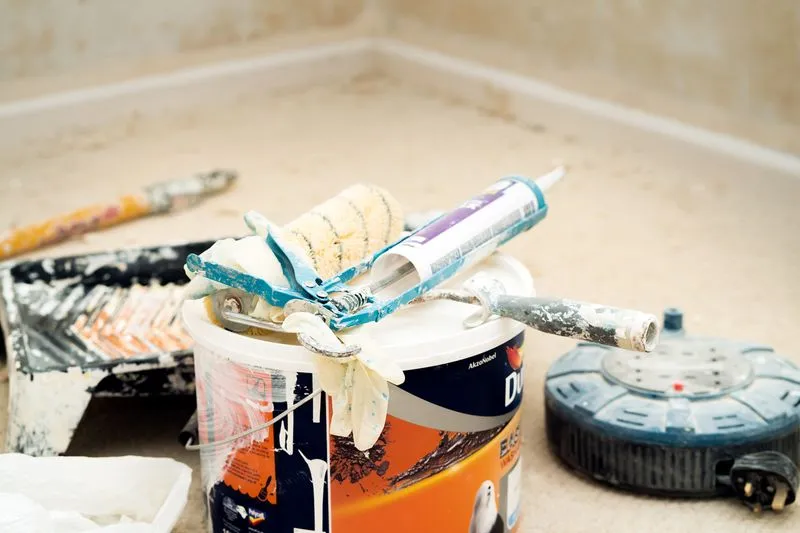Pot de peinture entamé avec rouleau, pistolet à mastic et gants sur le couvercle, bac et rallonge électrique au sol