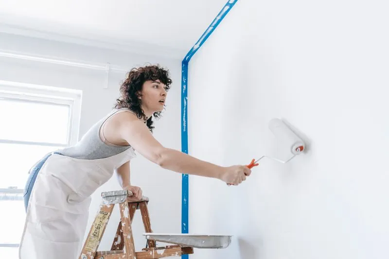Femme appuyée sur un escabeau qui passe le rouleau sur un mur blanc, angle protégé par un ruban de masquage bleu