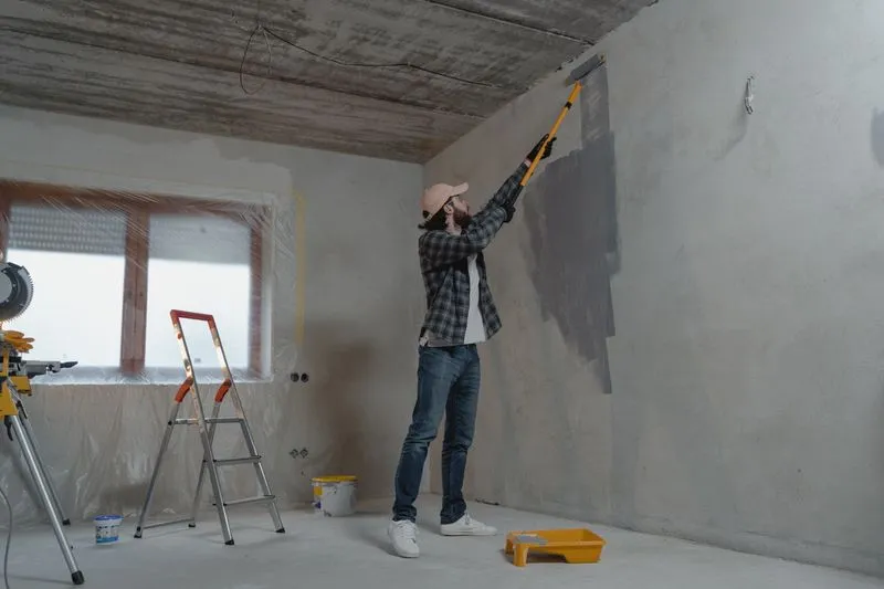 Pièce vidée en travaux, fenêtre entièrement couverte d'un film plastique, homme qui peint le haut du mur au rouleau à manche