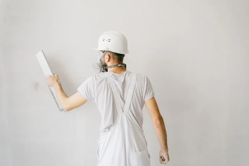 Artisan en salopette blanche, casque et masque respiratoire, qui lisse un mur blanc avec un large couteau à enduire