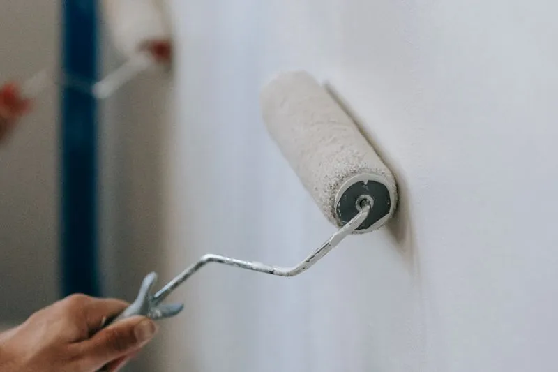 Main qui passe un rouleau chargé de peinture blanche sur un mur, ruban de masquage bleu dans l'angle