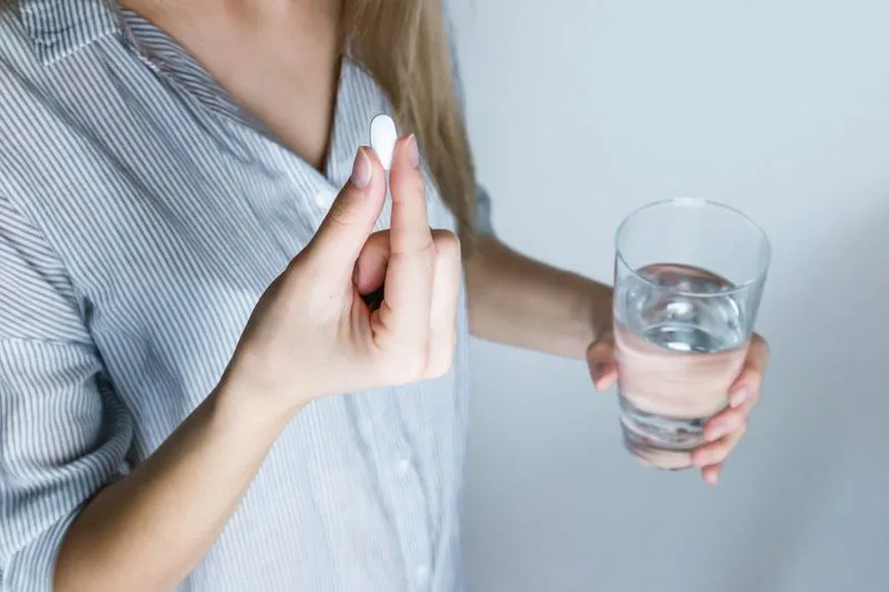Jeune femme qui tient un comprimé blanc entre ses doigts et un verre d'eau dans l'autre main
