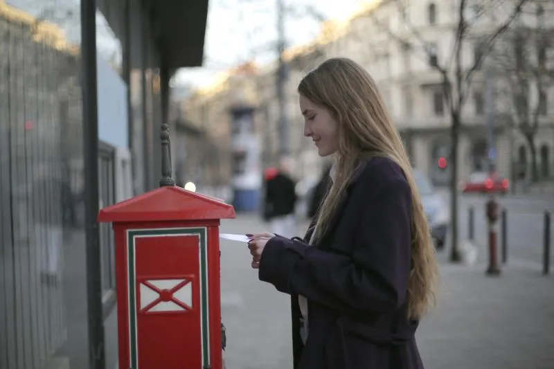 Jeune femme en manteau sombre qui poste une lettre dans une boîte aux lettres rouge, dans une rue en fin de journée