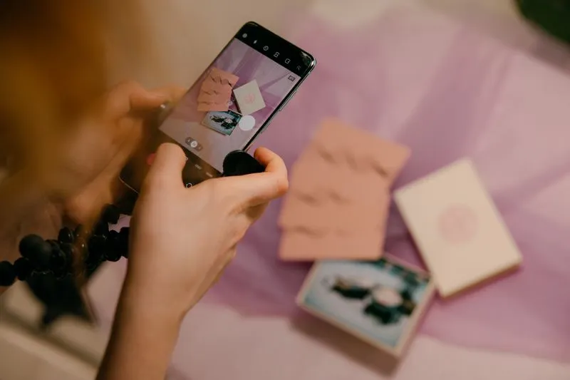 Mains qui photographient au smartphone des cartes et des enveloppes roses posées sur un tissu lilas