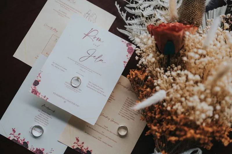 Faire-part de mariage et ses cartes assorties, avec des alliances posées dessus, à côté d'un bouquet de fleurs séchées