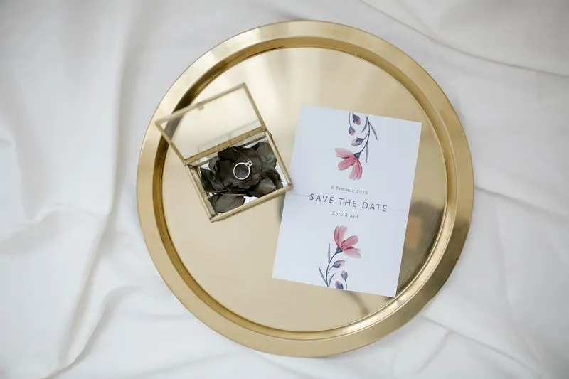 Carte save-the-date à fleurs roses sur un plateau doré, à côté d'une bague dans un écrin de verre