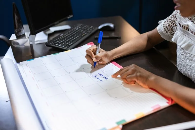 Main qui inscrit une date au stylo bleu sur un grand calendrier mensuel posé sur un bureau