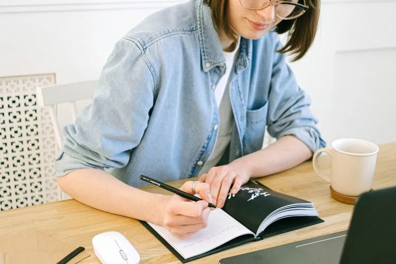 Jeune femme qui écrit dans un carnet ouvert sur un bureau en bois, à côté d'une tasse