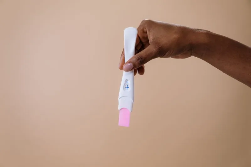 Main qui tient un test de grossesse dont la fenêtre affiche un signe plus, sur fond beige
