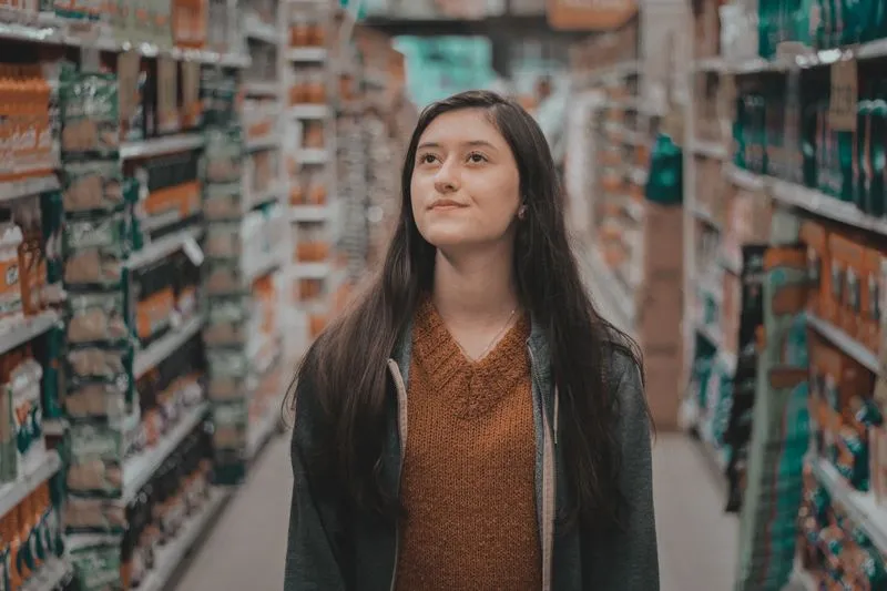 Jeune femme pensive, debout au milieu d'une allée de magasin bordée de rayonnages