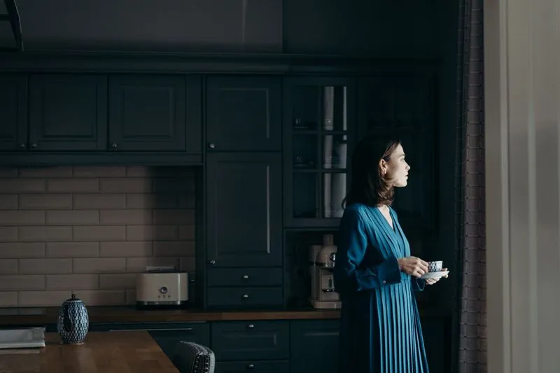 Femme debout dans une cuisine sombre, une tasse à la main, le regard tourné vers la fenêtre