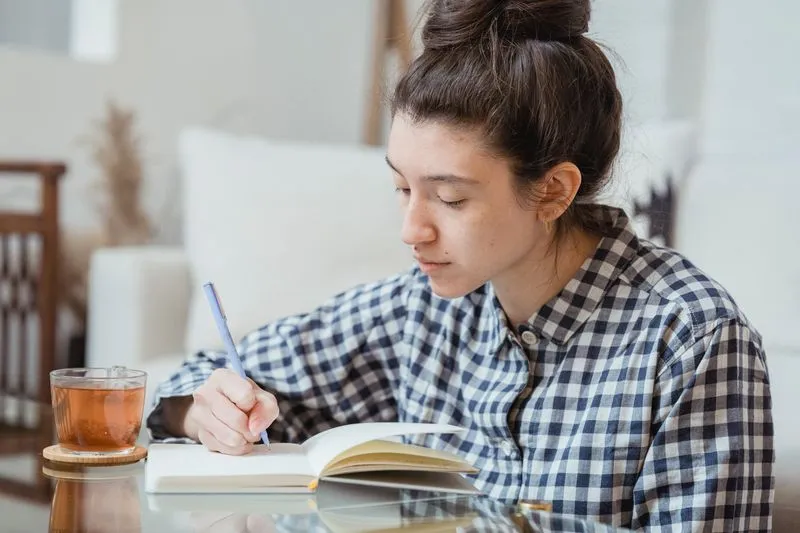 Jeune femme en chemise à carreaux qui écrit dans un carnet ouvert, un verre de thé posé à côté