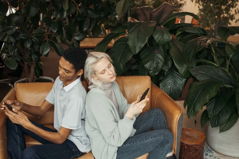 Jeune couple assis dos à dos dans un fauteuil en cuir, chacun absorbé par son téléphone, entouré de grandes plantes vertes