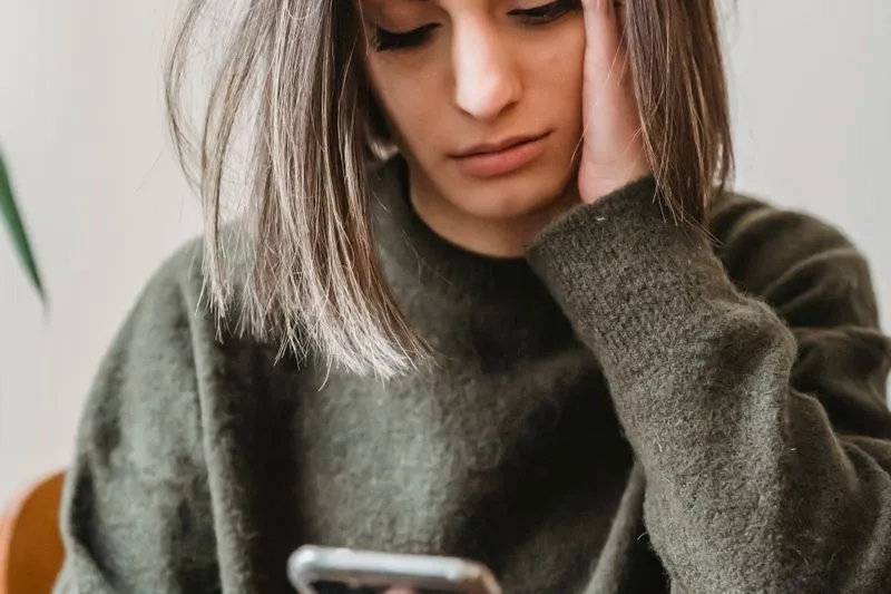 Jeune femme en pull kaki, la tête appuyée sur la main, les yeux baissés sur son téléphone, l'air soucieux