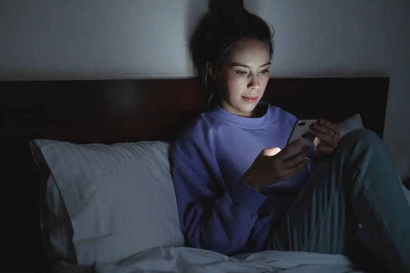 Jeune femme assise dans son lit la nuit, le visage éclairé par l'écran de son téléphone