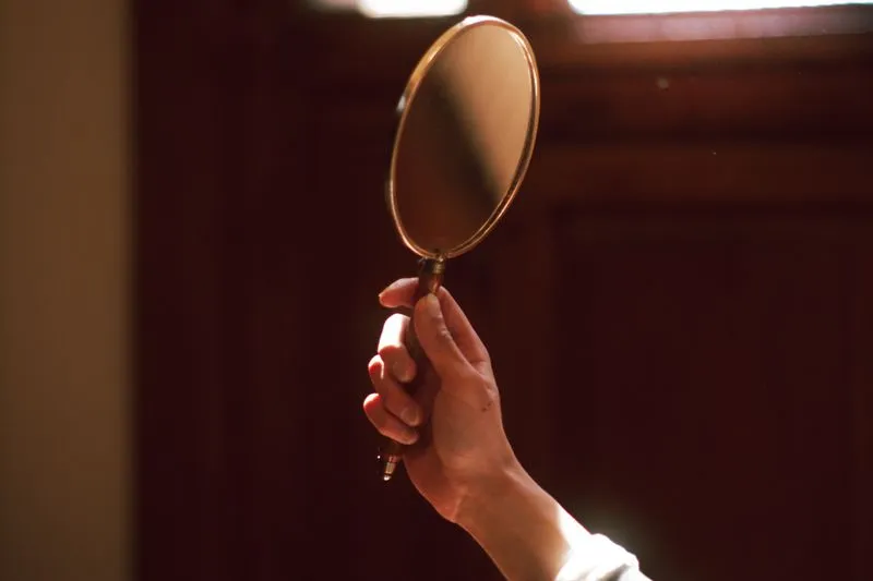 Main qui tient un miroir ovale cerclé de laiton, dans une pièce baignée de lumière chaude