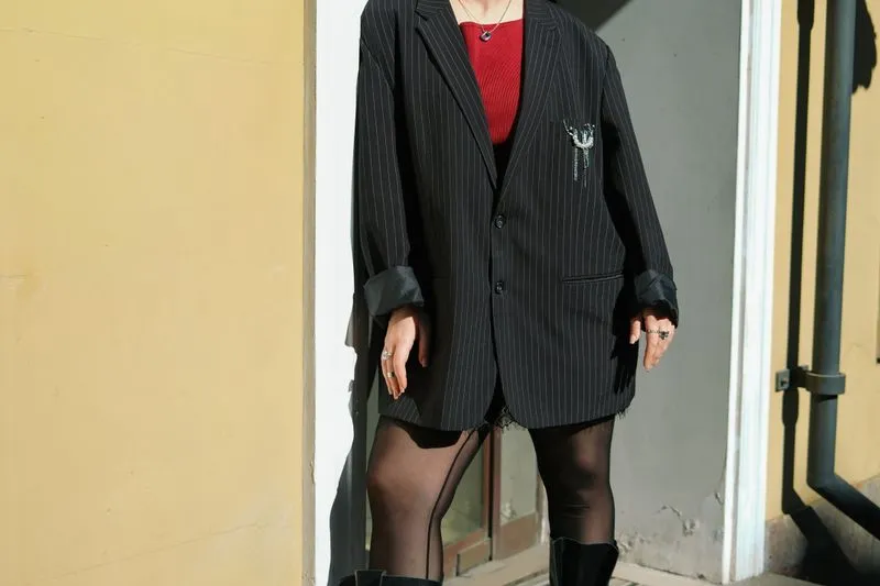 Blazer noir à fines rayures porté ample, manches retroussées, sur un haut rouge, des collants noirs et des bottes