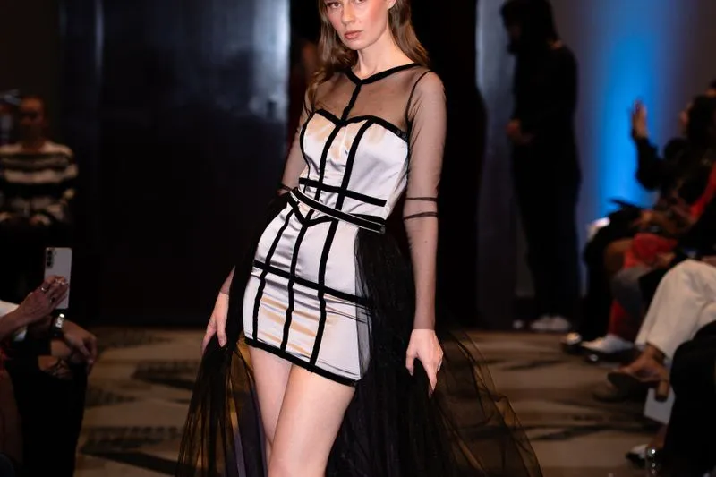 Mannequin en robe courte de satin gris à bandes noires et surjupe de tulle, sur un podium, devant des invités assis au premier rang