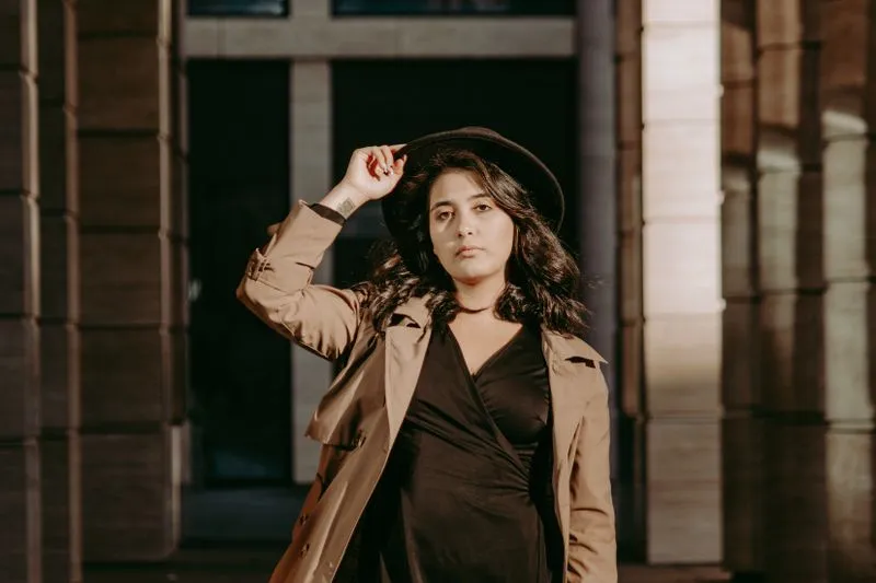 Femme en trench beige ouvert sur une robe portefeuille noire, chapeau noir sur la tête, entre des colonnes de pierre