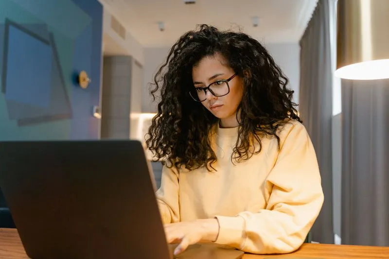 Femme aux cheveux bouclés et à lunettes, en sweat jaune pâle, qui tape sur un ordinateur portable