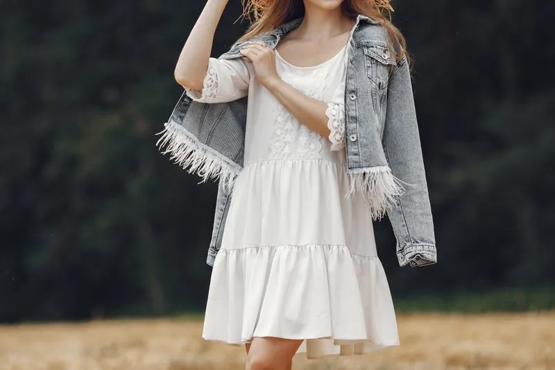 Femme en robe blanche courte à volants et manches de dentelle, une veste en jean à franges sur les épaules, dans un champ